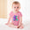 Baby in pink onesie with colorful cartoon bear, seated on white bed, light background.